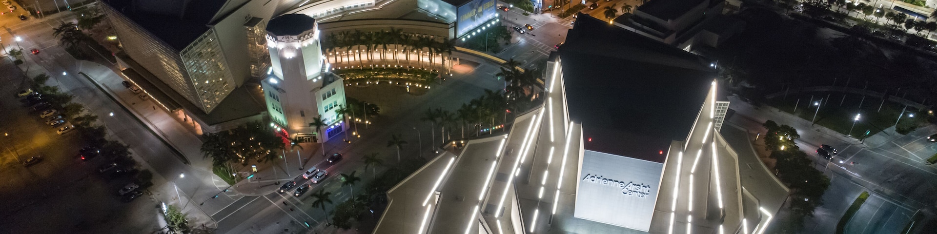 Un paisaje urbano nocturno con edificios iluminados, una plaza circular destacada y una gran estructura moderna con un diseño de techo singular.