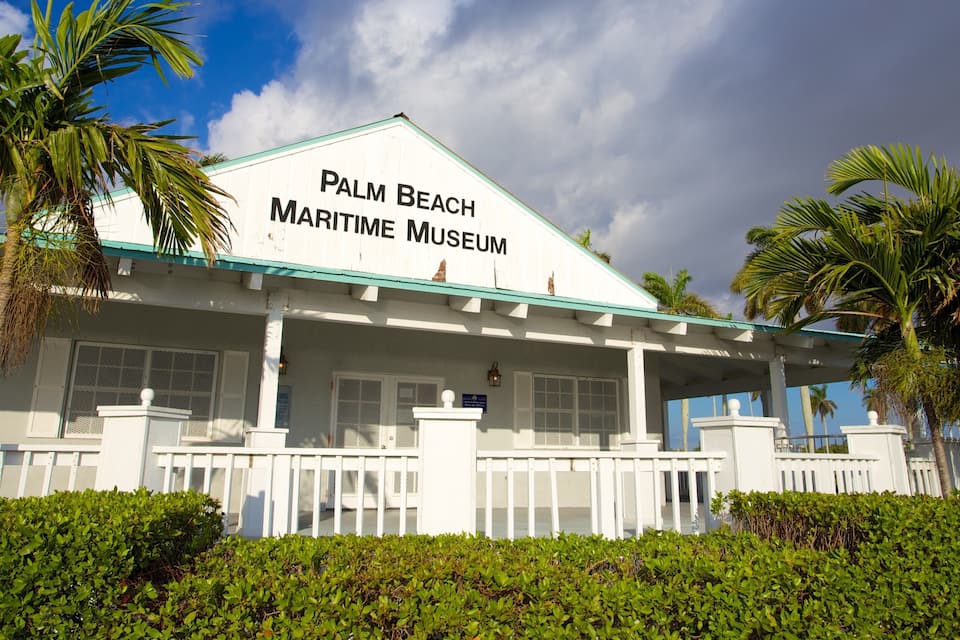 The Palm Beach Maritime Museum building with a white facade and green roof, surrounded by palm trees and greenery.