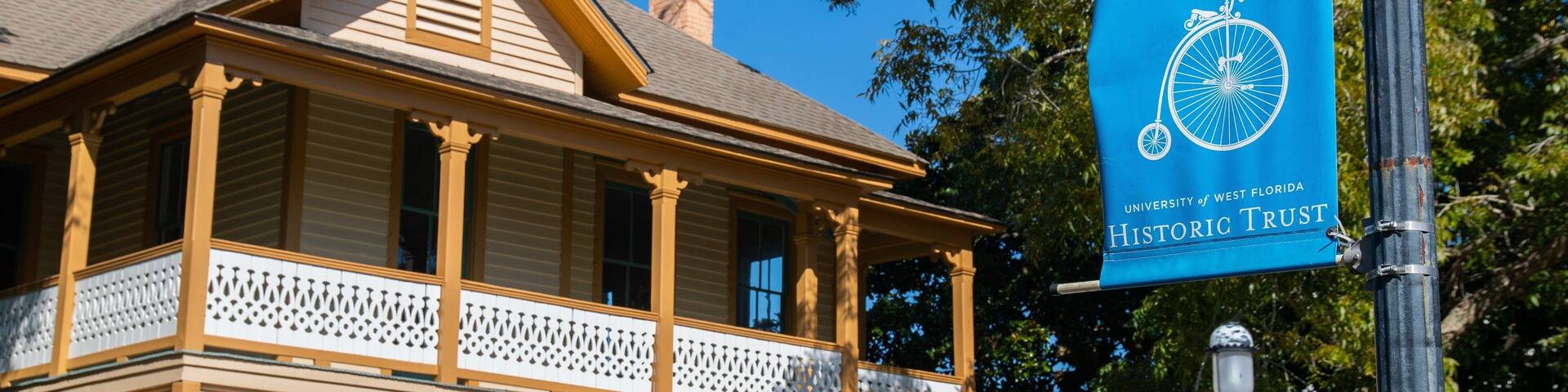 A historic building with a porch and columns, a blue banner with the Historic Pensacola Trust logo, and an American flag.