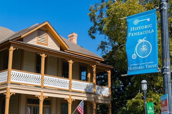 Un edificio histórico con un porche y columnas, un cartel azul con el logotipo de Historic Pensacola Trust y una bandera estadounidense.