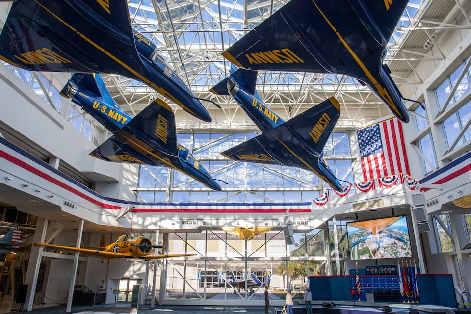 Interior de museu com aeronaves da Marinha dos EUA, bandeiras americanas e teto de vidro.