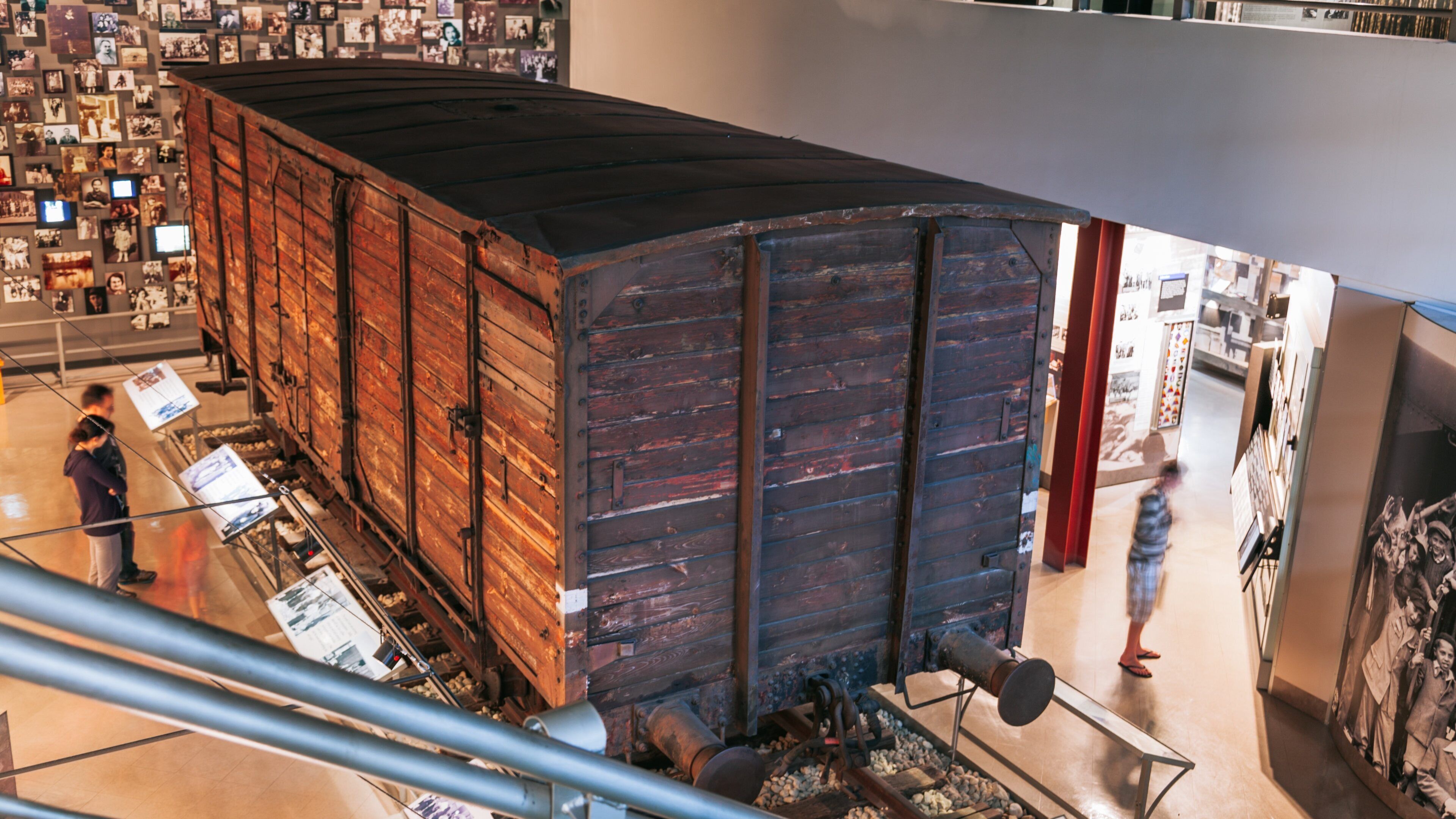 Un grand wagon de train en bois est exposé à l’intérieur, sous un toit en métal, et est entouré de photographies accrochées aux murs.