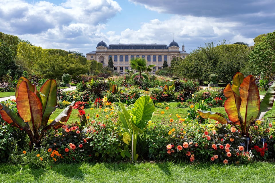 Un grand bâtiment doté d’un dôme et de colonnes, entouré d’un jardin bien entretenu où poussent de nombreuses fleurs aux couleurs vives et de grandes plantes tropicales.
