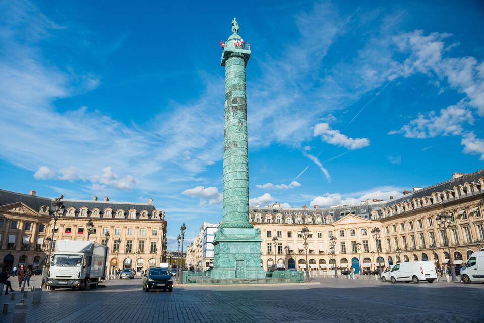A tall, green monument in a square with historic buildings around it.