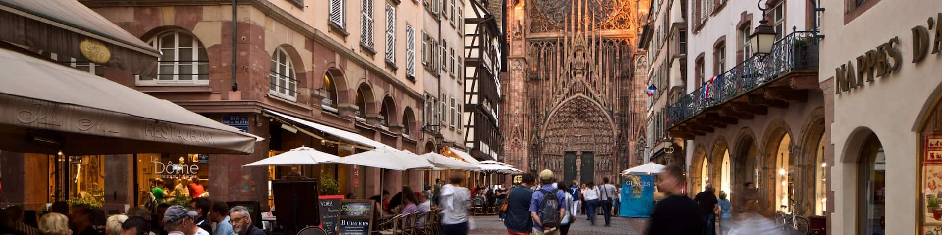 A cobblestone street with historic buildings, outdoor cafes, and a cathedral in the background.