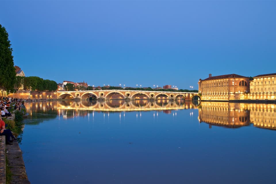 Uma ponte histórica sobre um rio calmo, com reflexo, cercada por edifícios e uma multidão de pessoas.