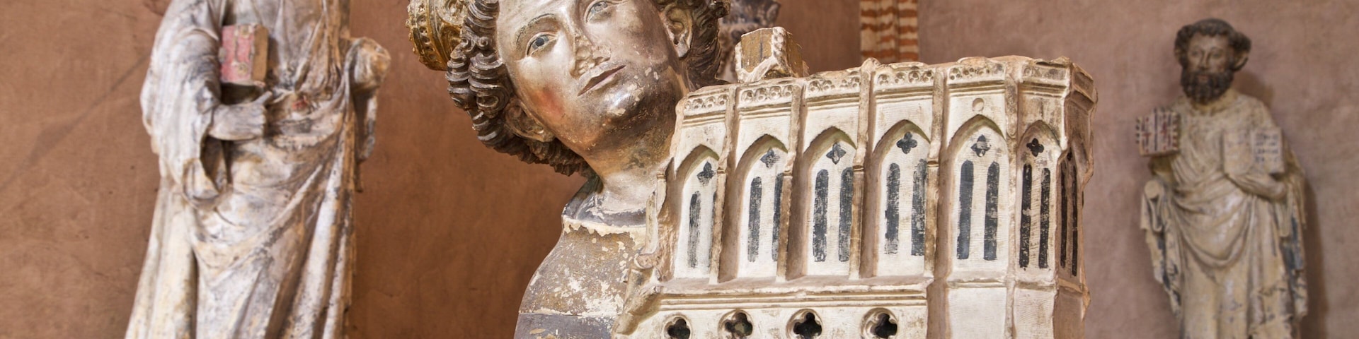 Uma estátua de uma figura vestida com manto, segurando um livro, com detalhes arquitetônicos góticos.