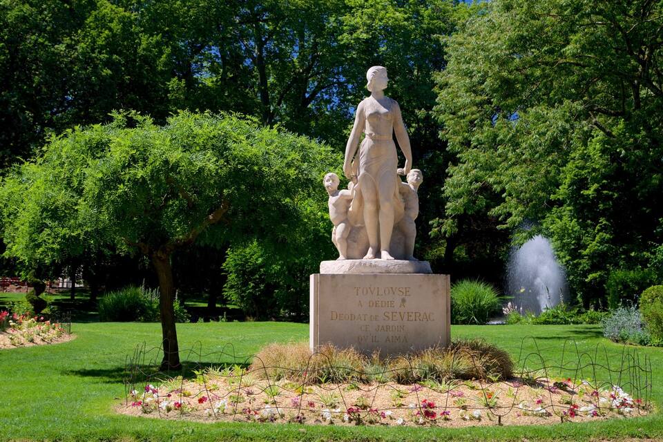 Una estatua de una mujer con un niño en un parque con abundante vegetación y una fuente.