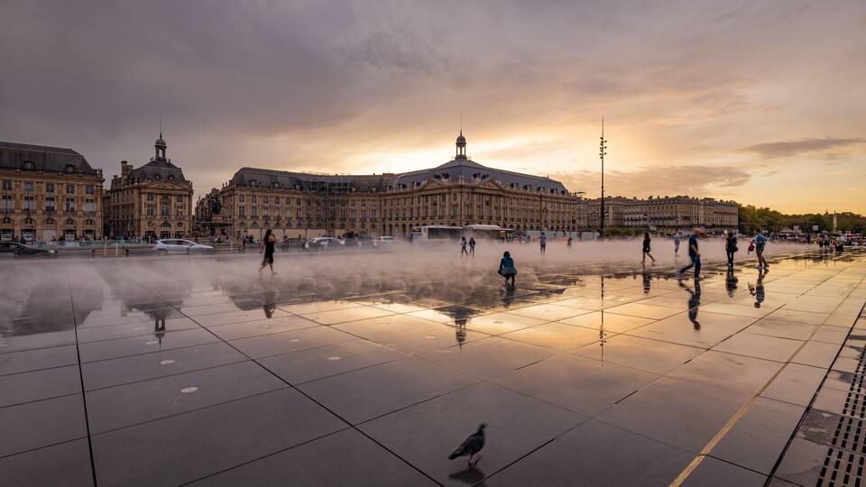 Una plaza histórica con una fuente, edificios que se reflejan y un atardecer.