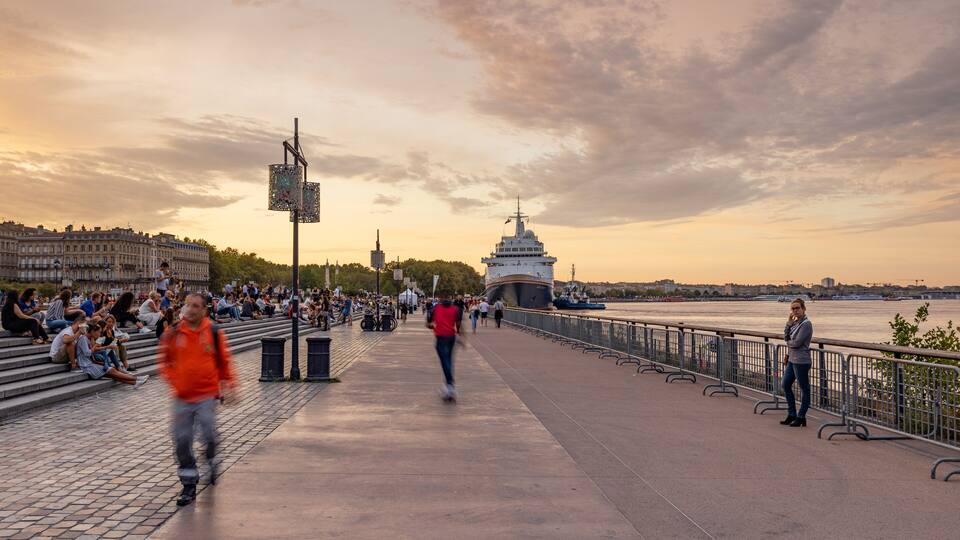 Un paseo fluvial con gente caminando y sentada, un barco grande amarrado y edificios históricos al fondo.