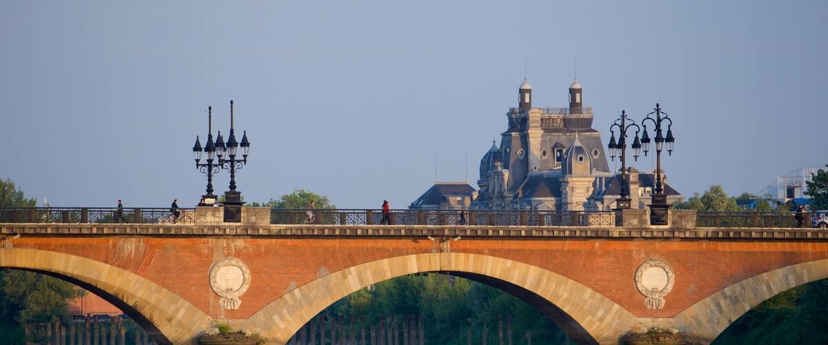 A stone bridge with arches over a river, a historic building with domes in the background, and street lamps along the bridge.