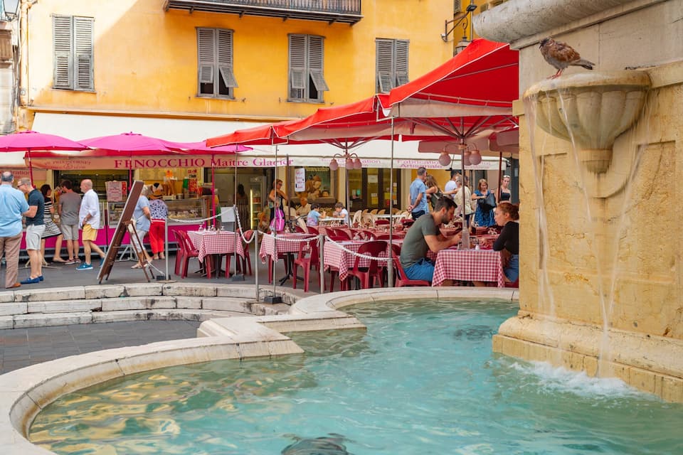 Una piazza pubblica con una fontana, posti a sedere all'aperto e un edificio giallo con persiane.