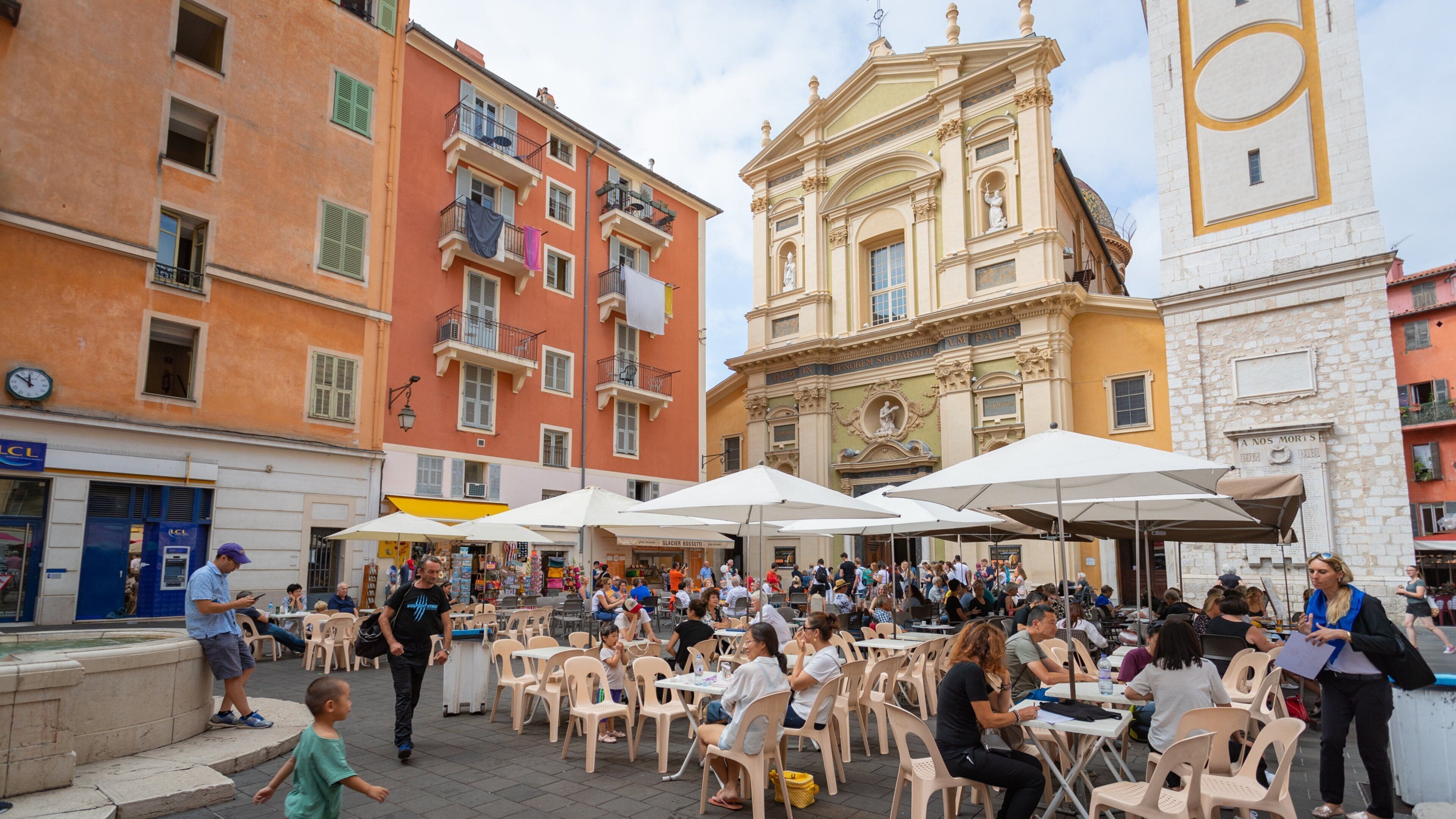 Un café al aire libre concurrido donde personas comen y conversan, con edificios históricos coloridos y una iglesia destacada de fondo.