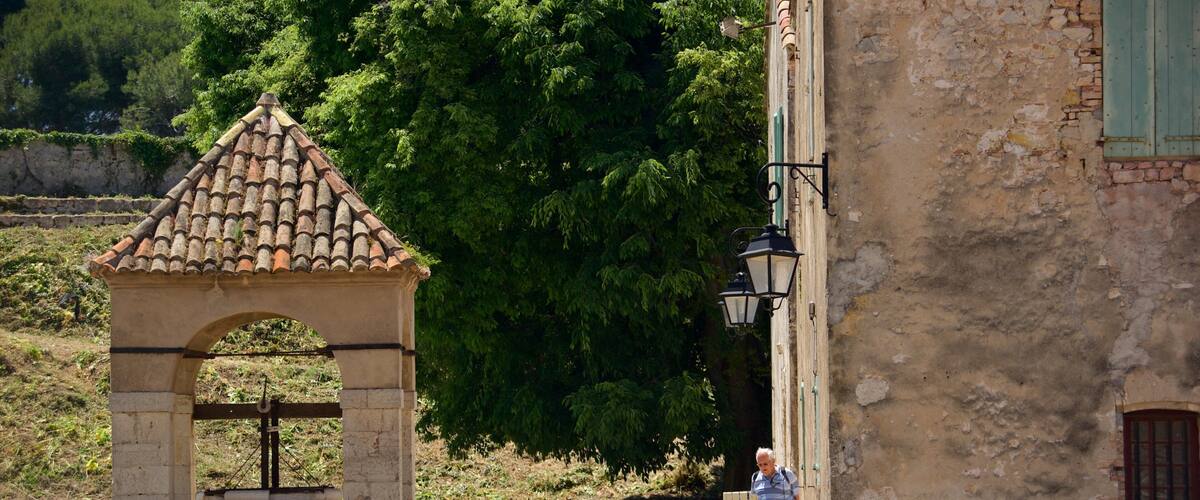 A stone archway with a tiled roof, a rustic building with a weathered facade, and a large tree in the background.