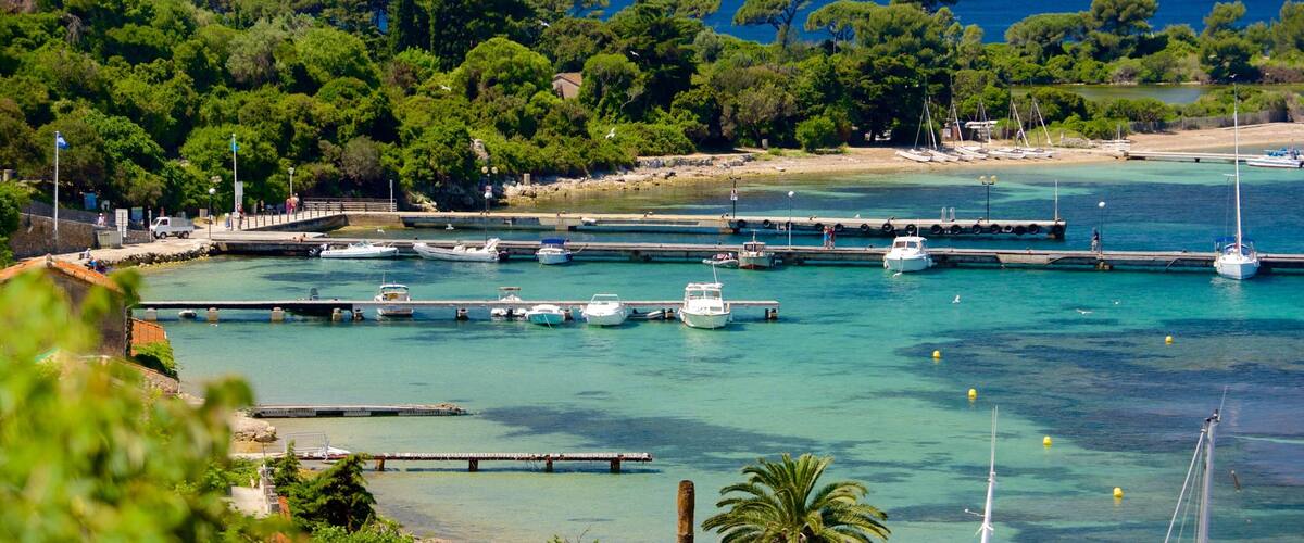 A coastal marina with boats docked, surrounded by lush greenery and a clear blue sea.
