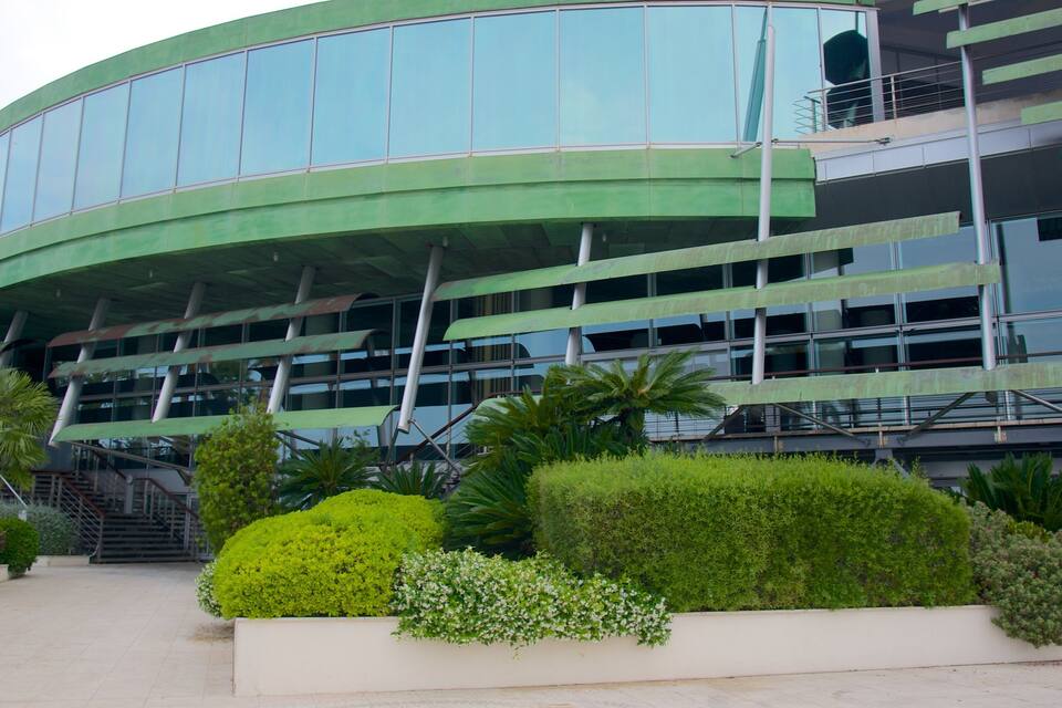 A modern building with curved glass windows and green awnings, surrounded by well-maintained bushes and palm trees.