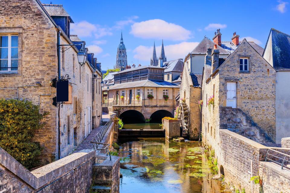 A narrow canal flanked by historic stone buildings, a bridge, and a prominent church spire in the background.