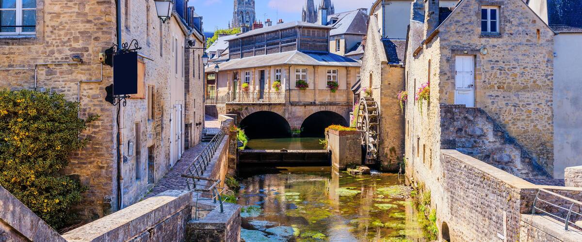 A narrow canal flanked by historic stone buildings, a bridge, and a prominent church spire in the background.