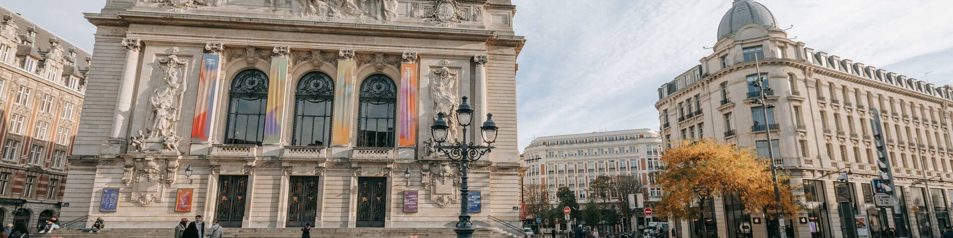 A historic building with ornate sculptures and columns, a cobblestone plaza, and a large tree with autumn leaves.