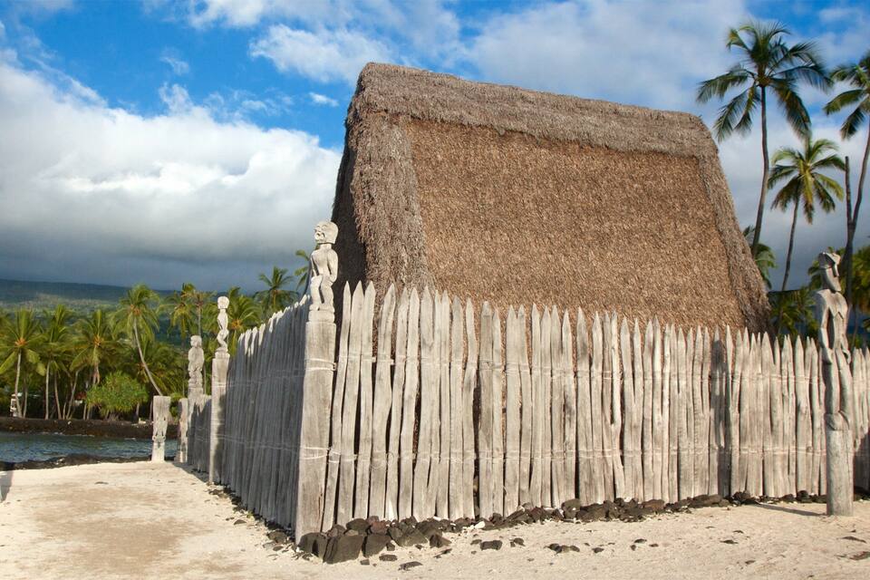 Eine traditionelle strohgedeckte Hütte mit Holzzaun, umgeben von Palmen und einem Strand.