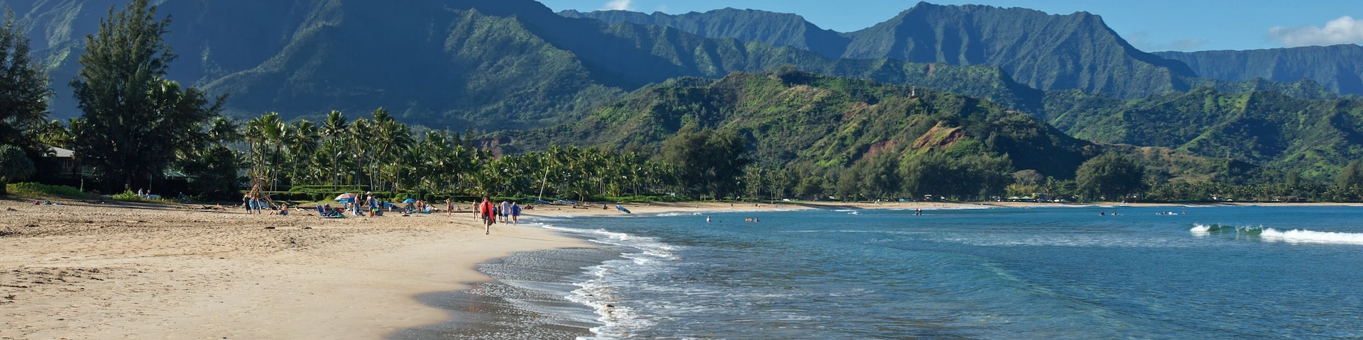 Uma praia com águas azuis cristalinas, areal arenoso e uma cordilheira ao fundo.