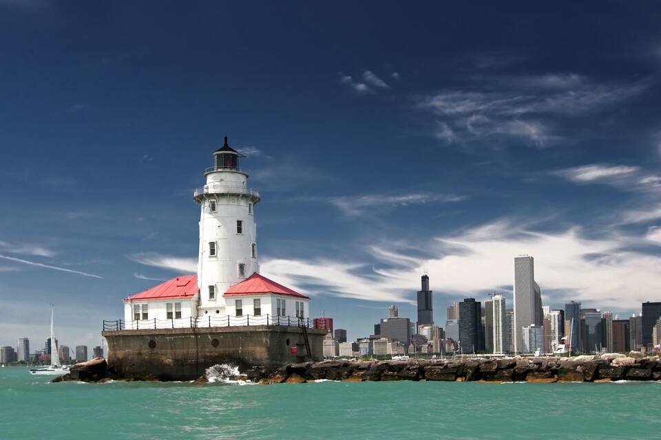 A lighthouse with a red roof, a city skyline in the background, and calm turquoise waters.