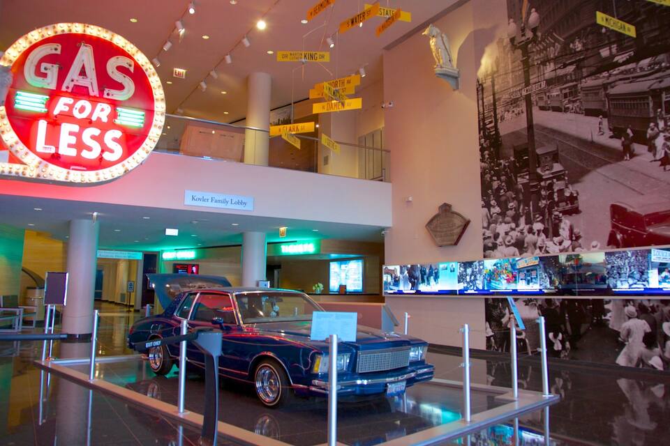 A vintage car in a museum exhibit, a neon sign reading "GAS FOR LESS", and a historical mural on the wall.
