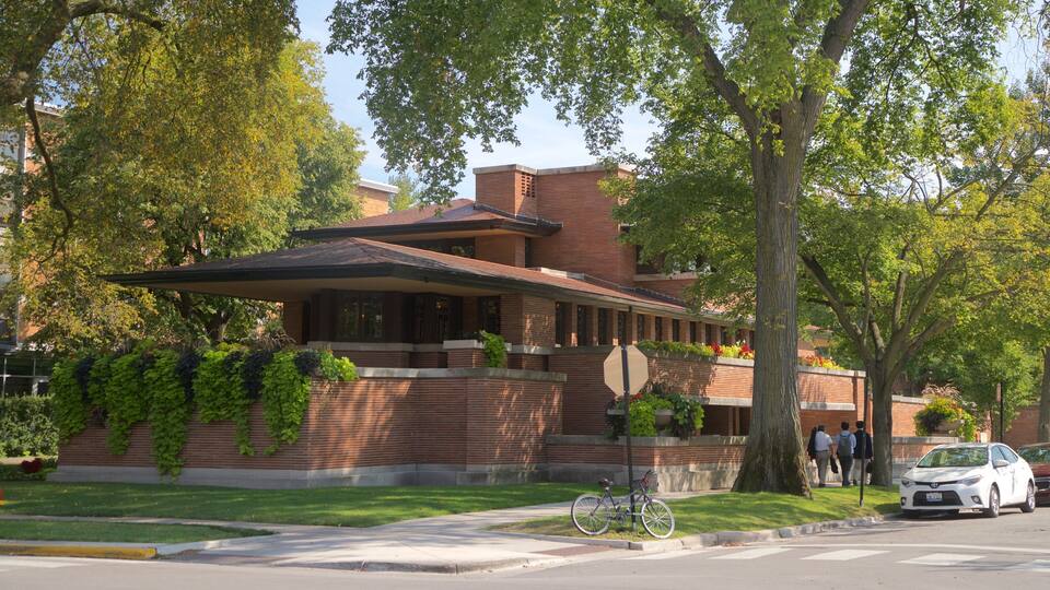 A modern brick house with a sloped roof and large windows, surrounded by lush green trees and a well-maintained lawn.