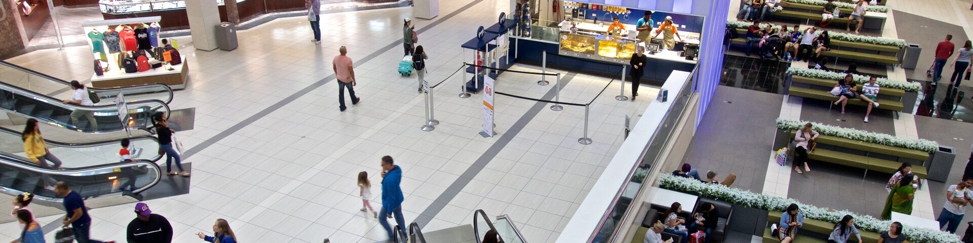 Un centro comercial con escaleras mecánicas, bancos y diversas tiendas, incluyendo Rogers & Holland's y una tienda de palomitas de maíz.