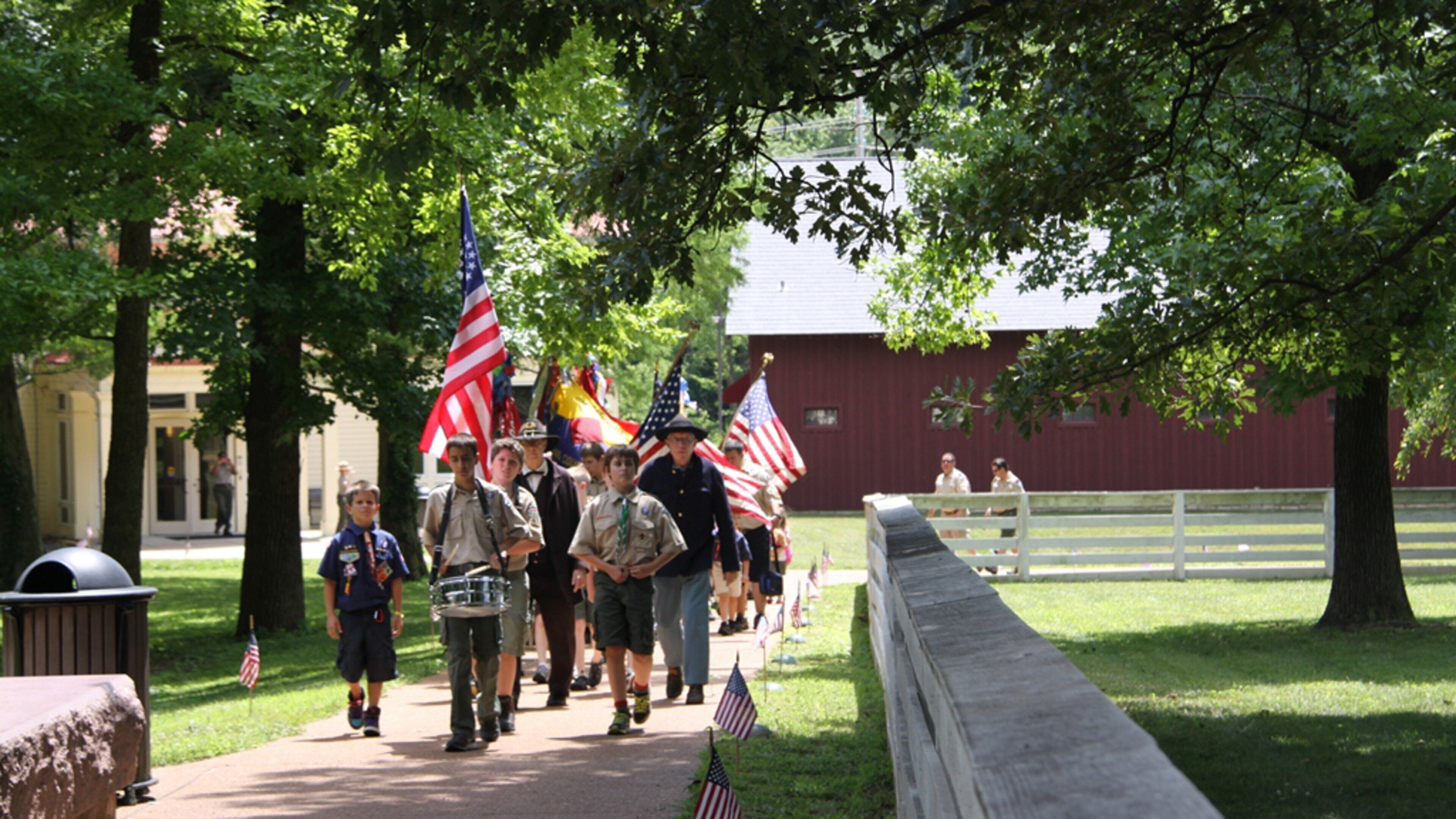 A group of Boy Scouts walking with American flags, a red barn, and a white fence in a park.