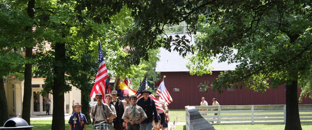 A group of Boy Scouts walking with American flags, a red barn, and a white fence in a park.