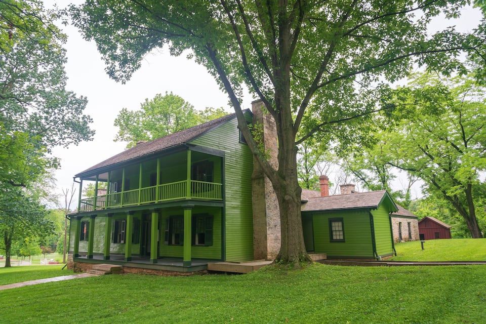 A green two-story house with a balcony, surrounded by lush trees and a well-maintained lawn.