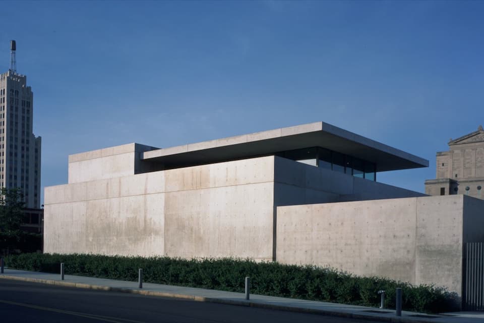Un bâtiment moderne en béton, doté d’un toit plat et de grandes fenêtres, entouré d’une haie et bordé par une rue.
