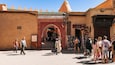 Una calle animada con un arco prominente con la inscripción 'MUSEE DEL MARRAKECH' que conduce a un edificio tradicional con techo de tejas verdes.