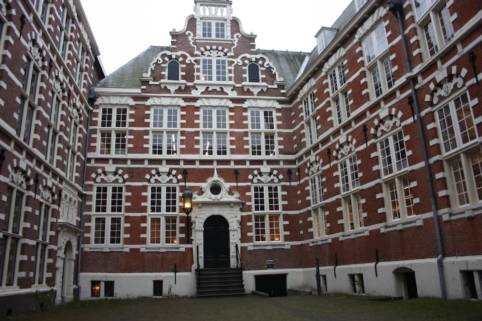 Un bâtiment historique aux murs de briques rouges et aux ornements blancs, doté de fenêtres décoratives et d’une entrée centrale.