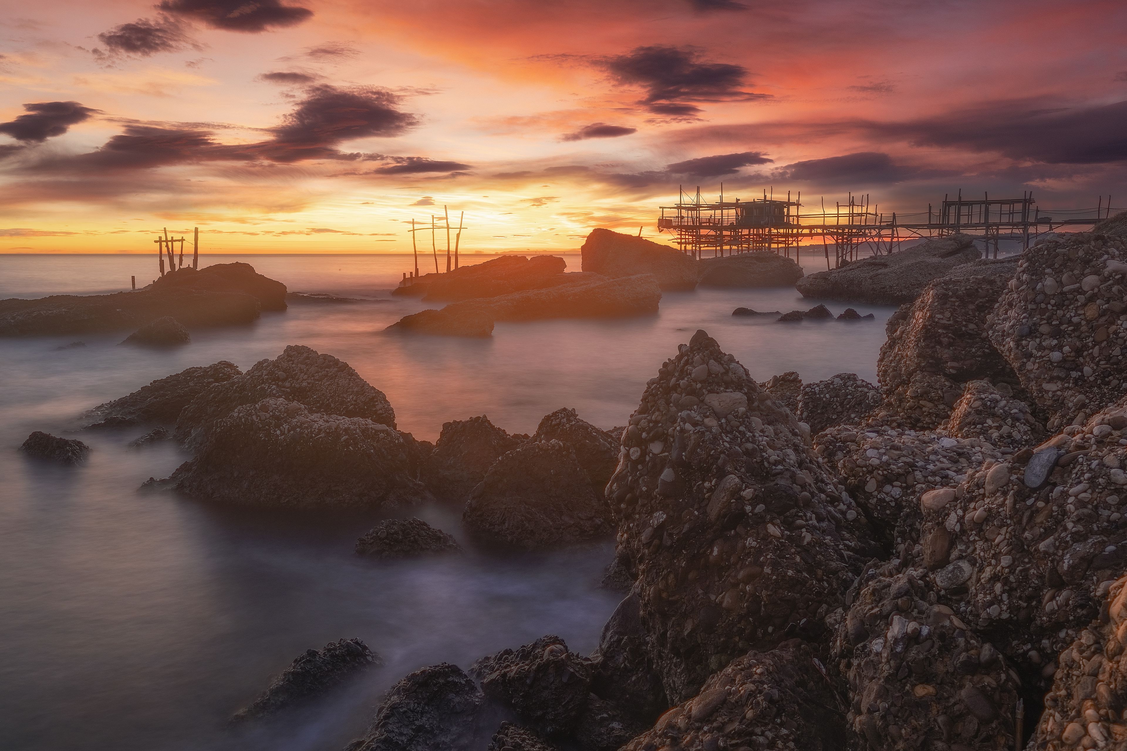 A rocky shoreline with a sunset, a pier with scaffolding, and calm water.