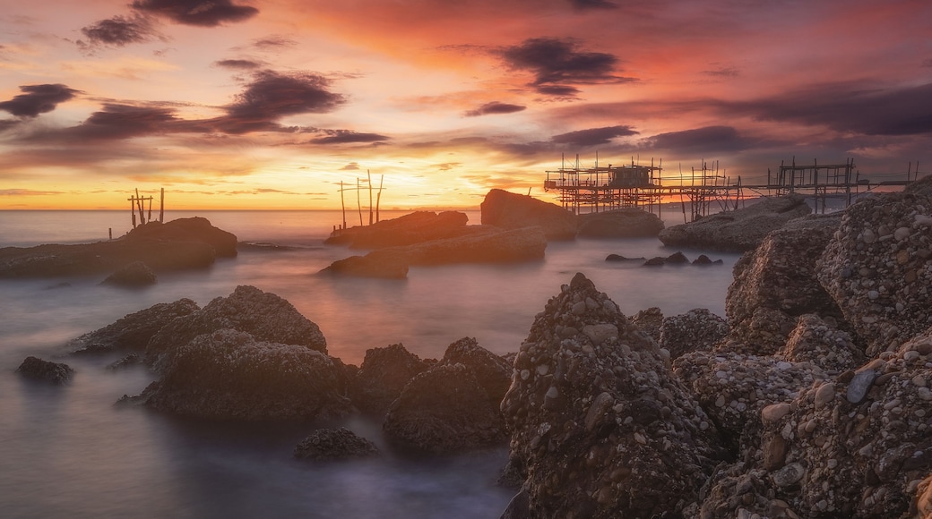 A rocky shoreline with a sunset, a pier with scaffolding, and calm water.