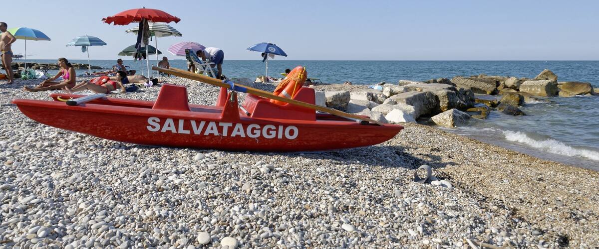 A red boat with 'SALVATAGGIO' written on it, a pebble beach, and people relaxing under umbrellas.
