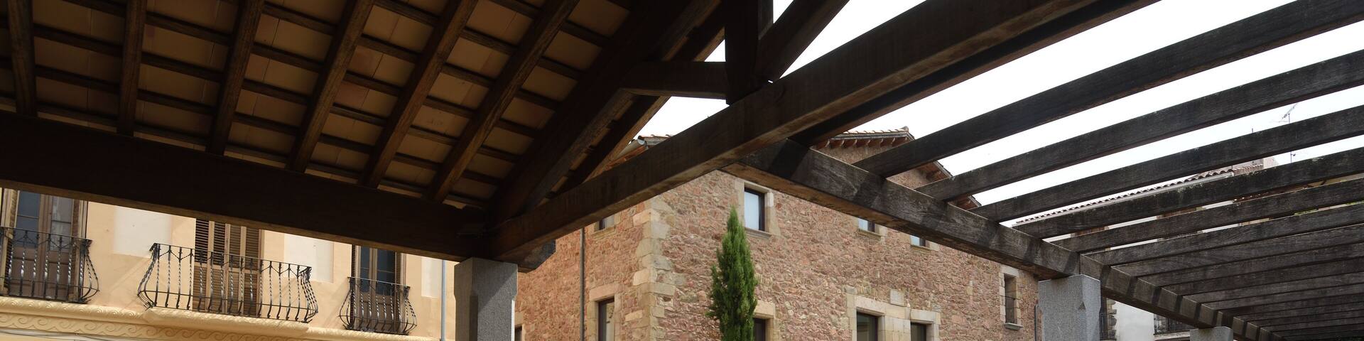 An open courtyard with a tiled floor, a wooden pergola, and a brick building with a stone facade.