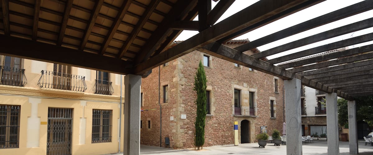 An open courtyard with a tiled floor, a wooden pergola, and a brick building with a stone facade.