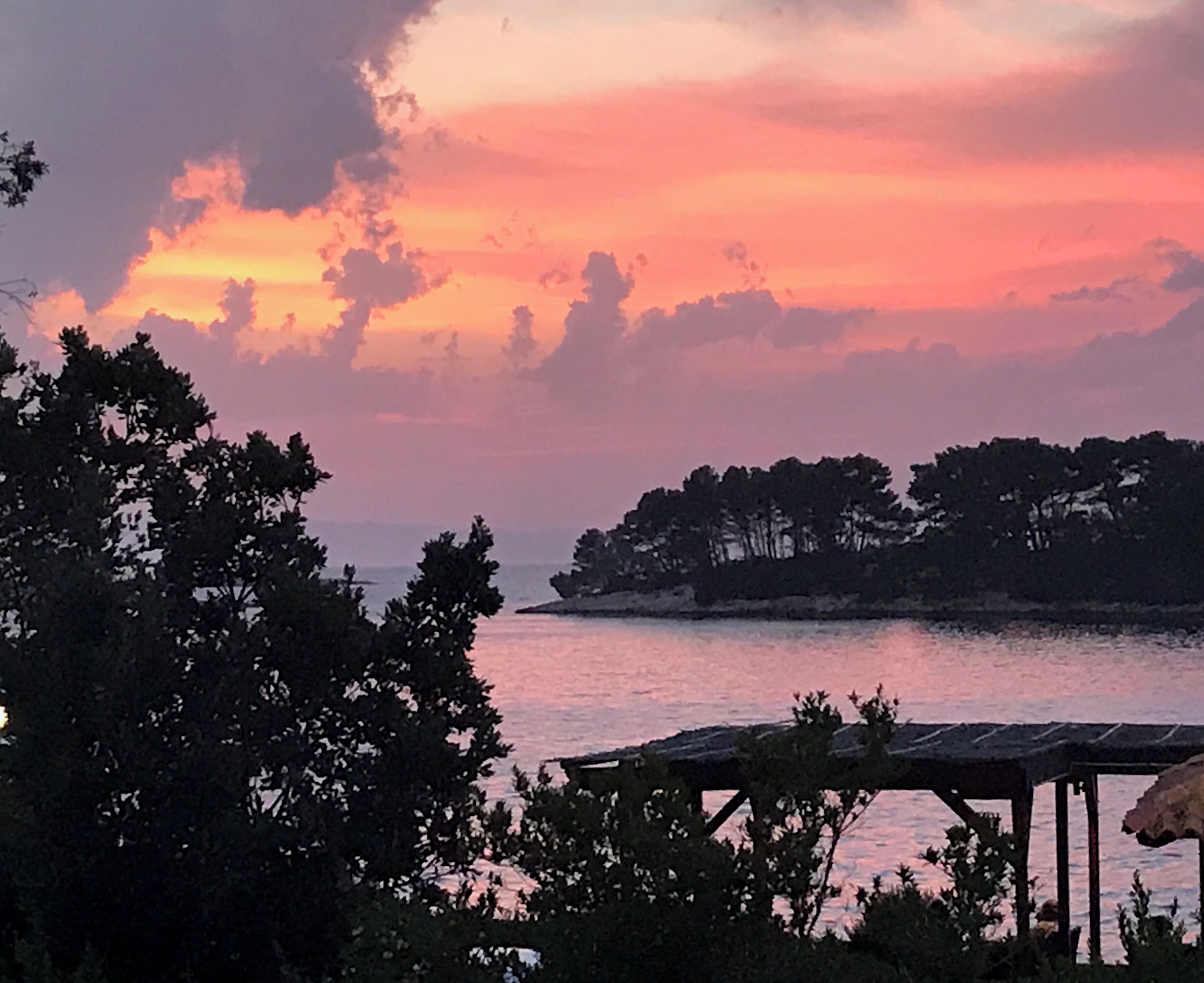 A sunset over a calm body of water with trees and a wooden structure in the foreground.