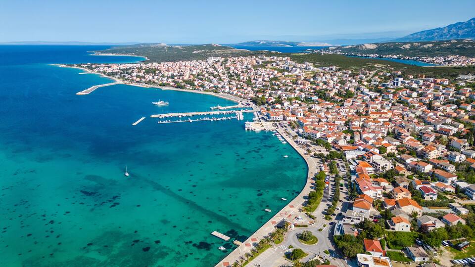 An aerial view of a coastal town with a marina, clear turquoise waters, and a hilly backdrop.