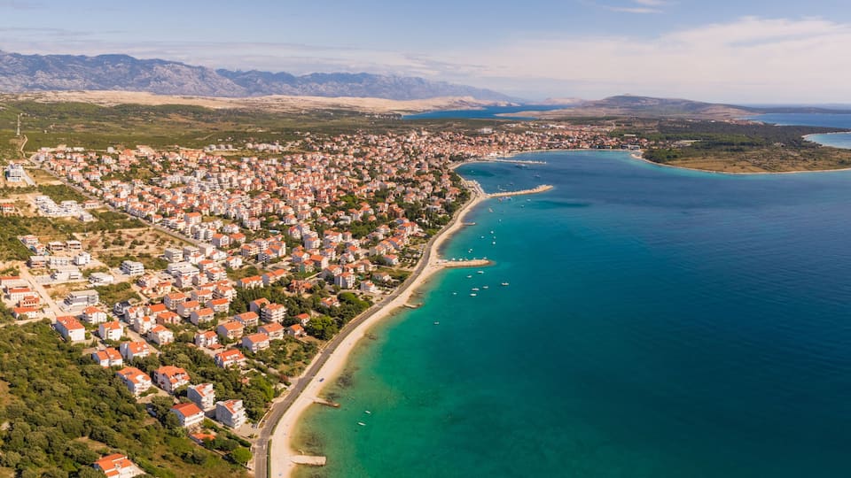 A coastal town with a long beachfront, clear turquoise waters, and a mountainous backdrop.