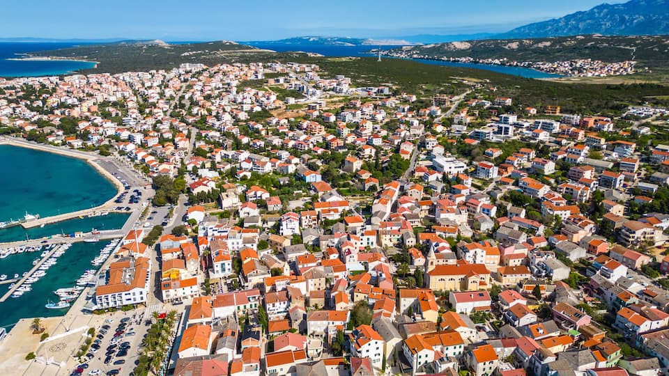 A coastal town with dense housing, a marina, and a clear view of the sea and mountains in the distance.