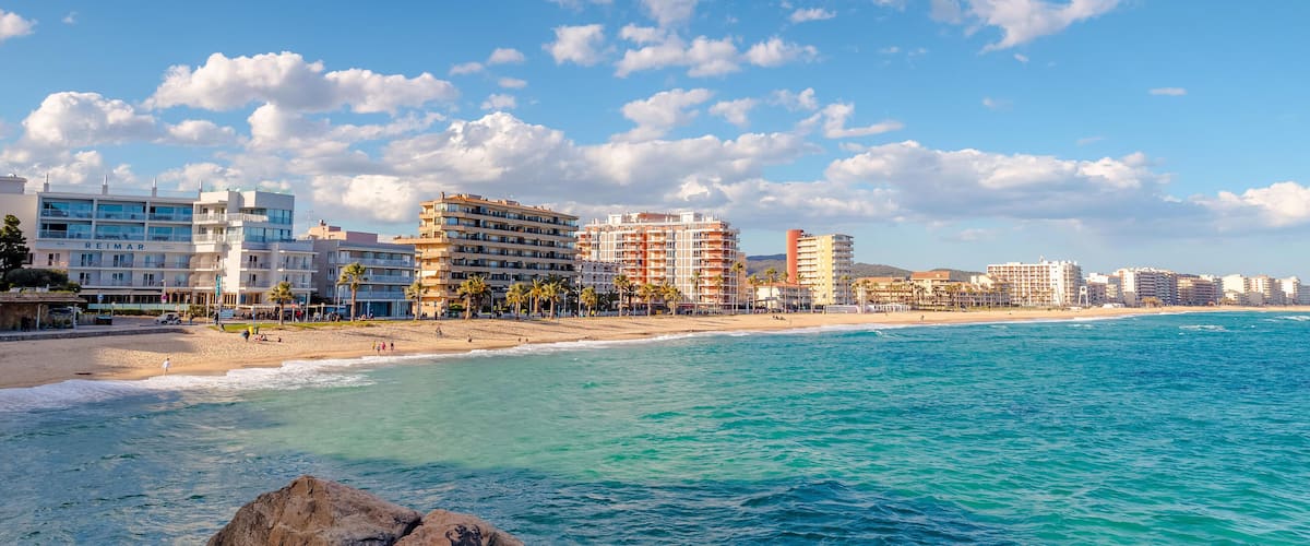 A coastal cityscape with a beach, clear blue waters, and a variety of buildings including a hotel named REIMAR.