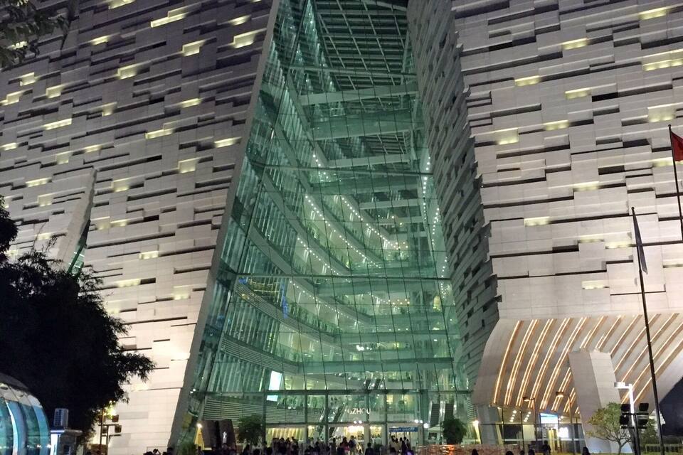 A modern glass building with a unique angular design, surrounded by a bustling urban environment at night.