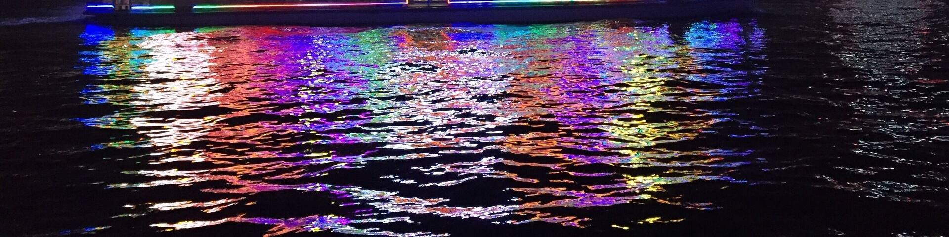 A brightly lit riverboat with traditional architecture, reflecting colorful lights on the water, set against a city skyline with modern buildings.