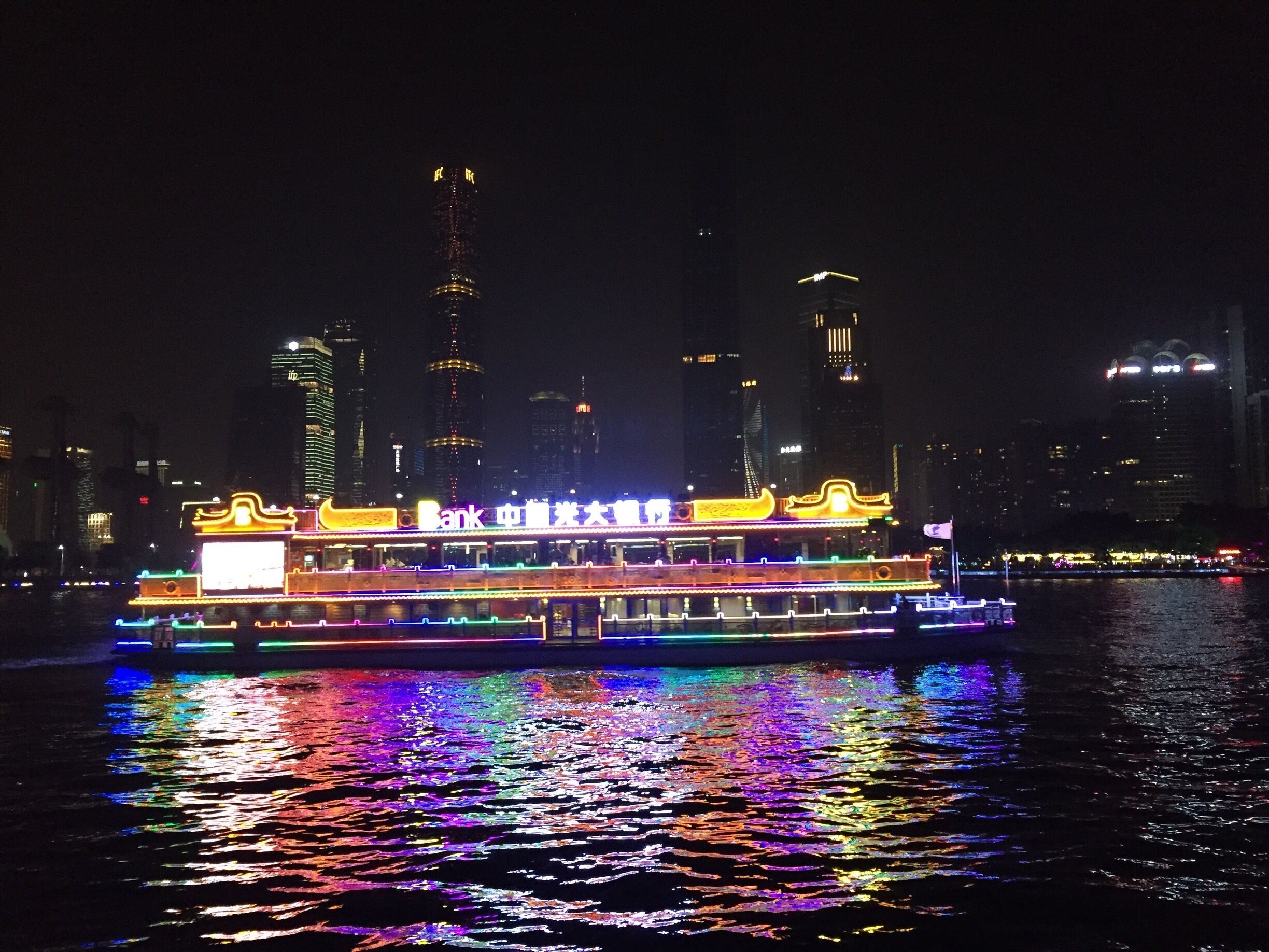 A brightly lit riverboat with traditional architecture, reflecting colorful lights on the water, set against a city skyline with modern buildings.