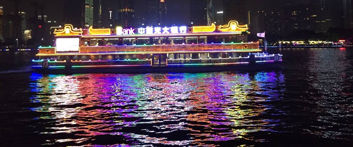 A brightly lit riverboat with traditional architecture, reflecting colorful lights on the water, set against a city skyline with modern buildings.