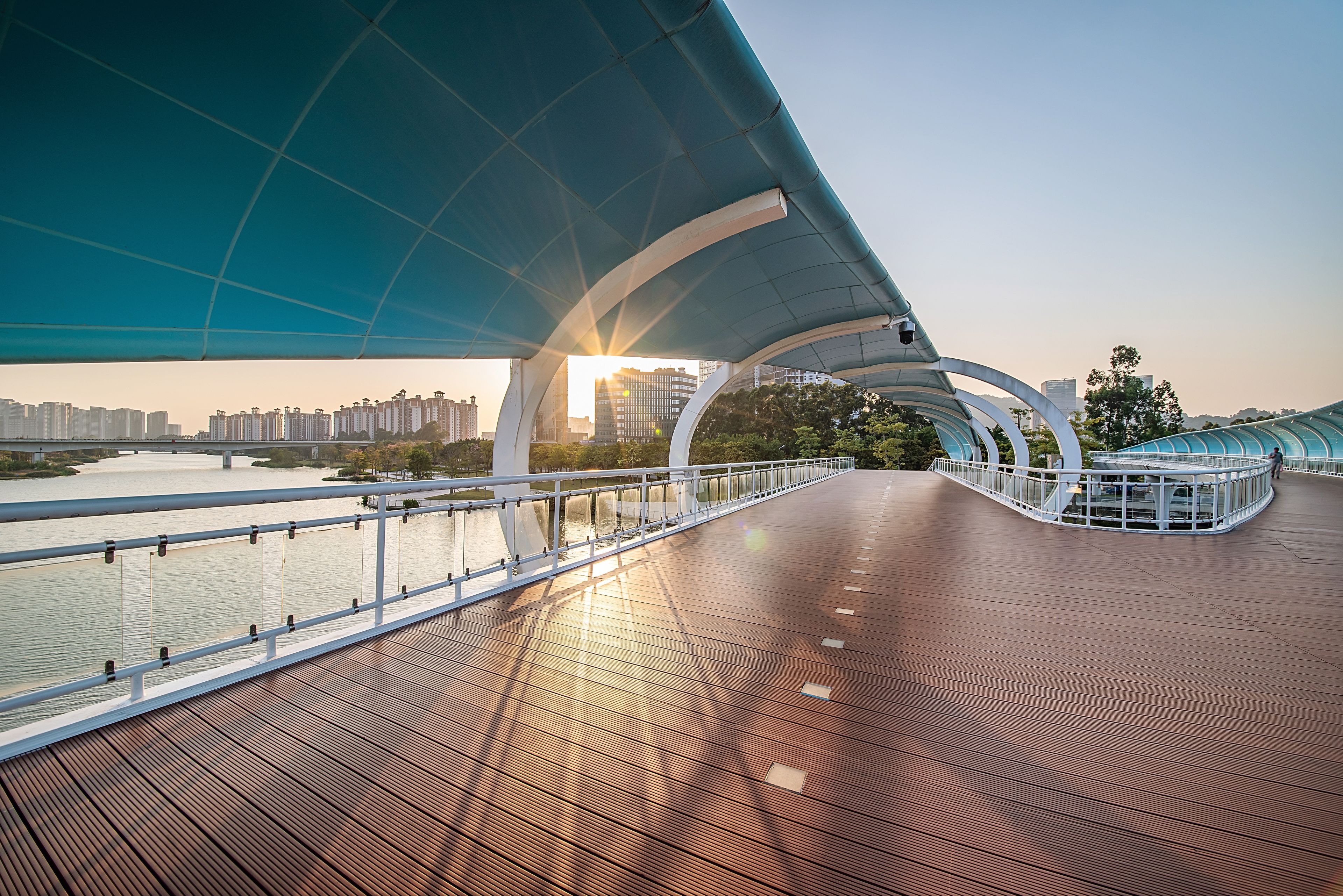 A modern bridge with a wooden deck and a blue canopy, overlooking a river with buildings in the background.